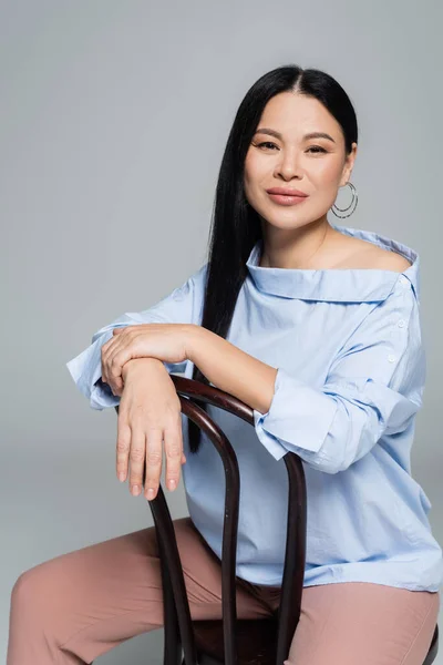 Щаслива азіатська жінка в блузці сидить на стільці ізольовано на сірому — стокове фото