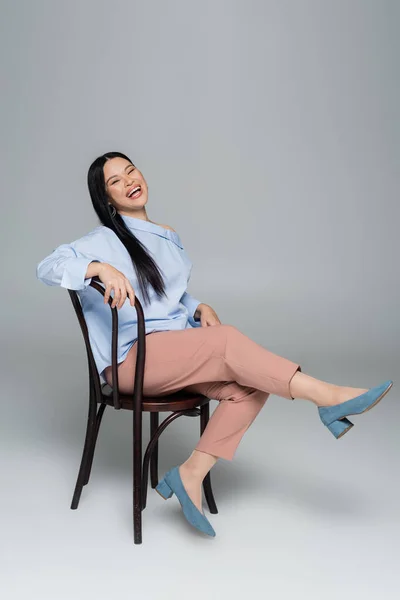 Щаслива і стильна азіатка позує на стільці на сірому фоні — стокове фото