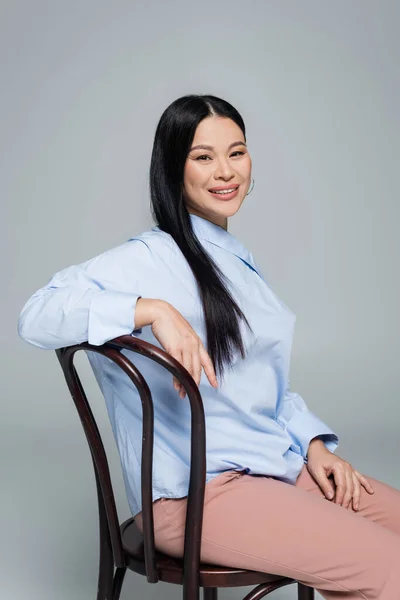 Positive asian woman in blouse sitting on chair and looking at camera isolated on grey — Stock Photo