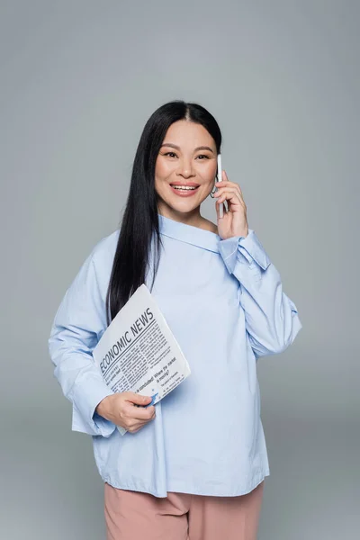 Улыбающаяся азиатка, держащая экономическую газету и разговаривающая на смартфоне изолированная на сером — стоковое фото