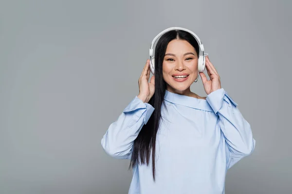 Fröhliche asiatische Frau in Bluse hört Musik in Kopfhörern isoliert auf grau — Stockfoto