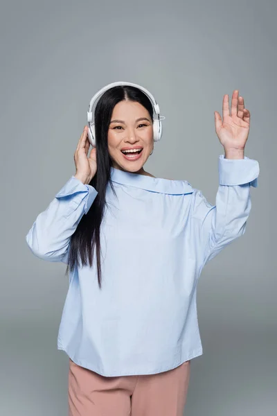 Positive asian woman in headphones waving hand isolated on grey — Stock Photo