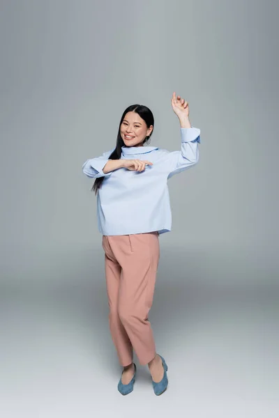 Улыбающаяся азиатская модель в блузке танцующая на сером фоне — стоковое фото