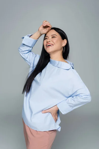 Positive asian woman in blue blouse holding hand on hip isolated on grey — Stock Photo
