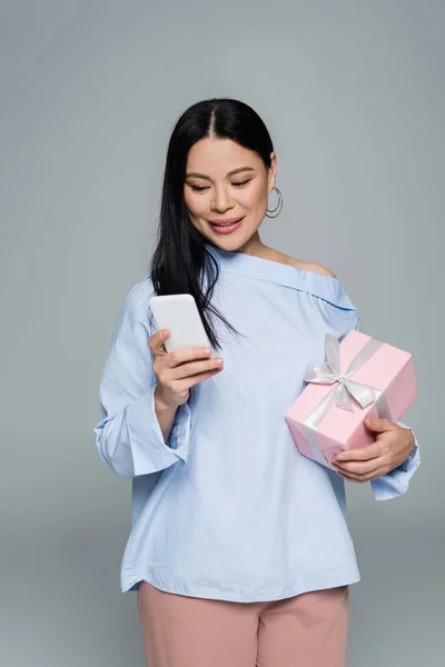Позитивная азиатка, использующая смартфон и держащая подарок, изолированный на сером — стоковое фото