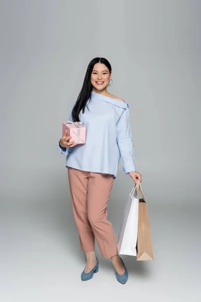 Cheerful asian woman holding gift and shopping bags on grey background — Stock Photo