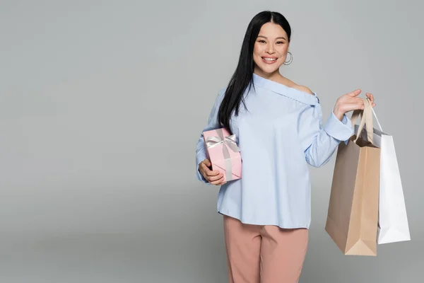 Smiling asian woman holding shopping bags and present isolated on grey — Stock Photo