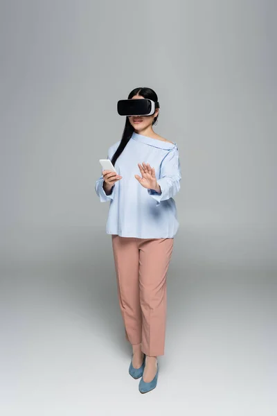 Trendy woman in vr headset using mobile phone on grey background — Stock Photo