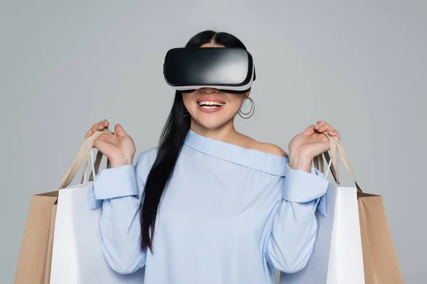 Allegro donna in auricolare vr che tiene borse della spesa isolate sul grigio — Foto stock