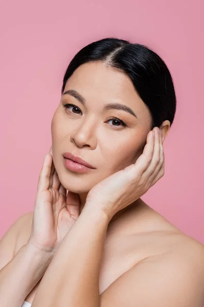 Modelo asiático con maquillaje tocando mejillas aisladas en rosa - foto de stock