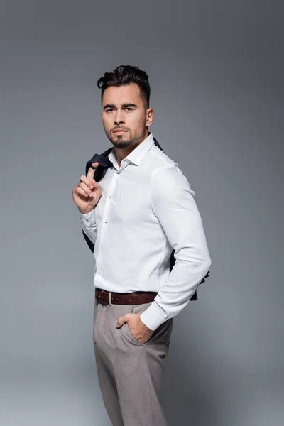 Joven hombre de negocios barbudo sosteniendo chaqueta y de pie con la mano en el bolsillo en gris - foto de stock