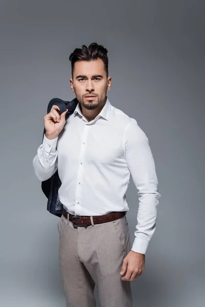 Joven hombre de negocios barbudo con camisa blanca sosteniendo chaqueta aislada en gris - foto de stock