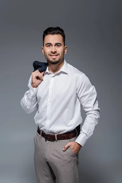 Hombre de negocios feliz sosteniendo chaqueta y de pie con la mano en el bolsillo aislado en gris - foto de stock