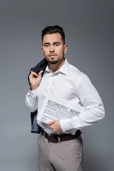 Young bearded businessman in suit holding newspaper and blazer isolated on grey — Stock Photo