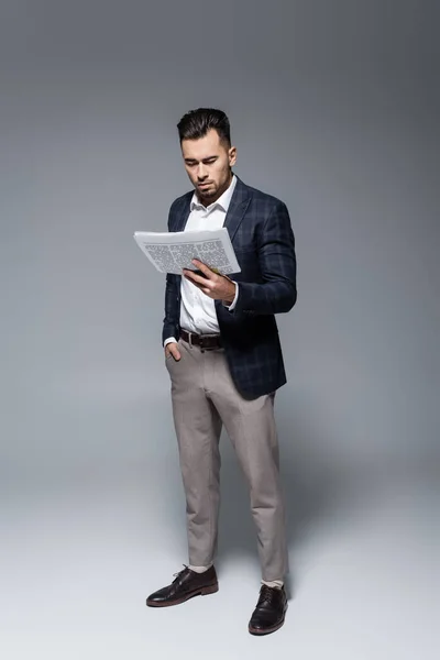 Intera lunghezza del giovane uomo d'affari barbuto in giacca a scacchi leggendo giornale su grigio — Foto stock