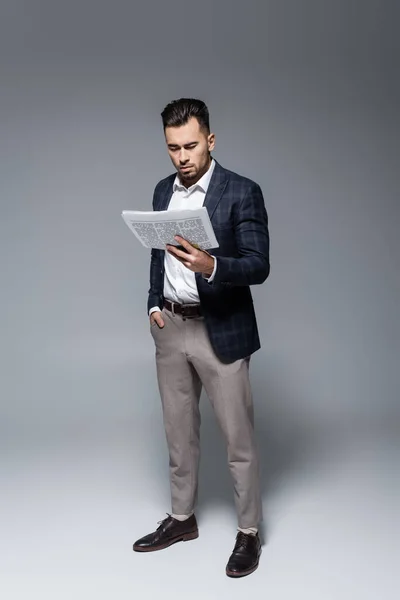 Longitud completa del joven hombre de negocios barbudo en el periódico de lectura de chaqueta a cuadros en gris — Stock Photo