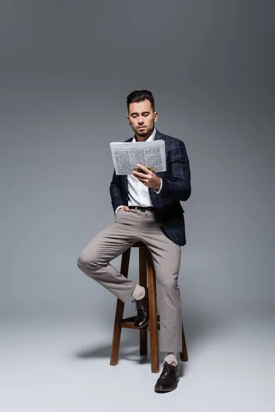 Longitud completa de hombre de negocios barbudo en chaqueta a cuadros sentado en la silla y leyendo el periódico en gris - foto de stock