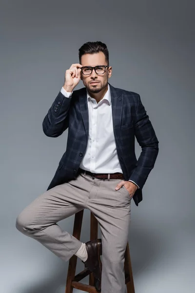 Homme d'affaires barbu en blazer ajuster les lunettes et assis sur la chaise tout en posant avec la main dans la poche isolé sur gris — Photo de stock