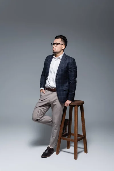 Vollbärtiger Geschäftsmann im Anzug steht mit der Hand in der Tasche in der Nähe von Holzstuhl auf grau — Stockfoto