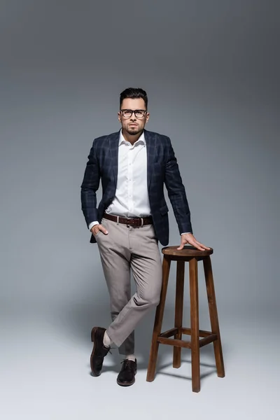 Homme d'affaires barbu en costume et lunettes debout avec la main dans la poche près de la chaise sur gris — Photo de stock