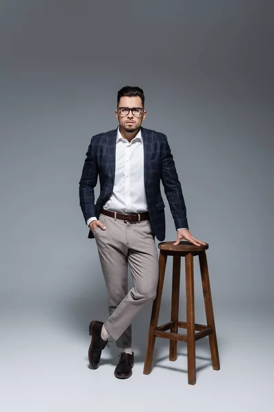 Full length of bearded businessman in suit and glasses standing with hand in pocket near chair on grey — Stock Photo