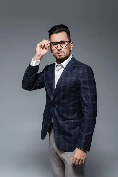 Hombre de negocios barbudo en blazer ajustando gafas y mirando a la cámara aislada en gris - foto de stock