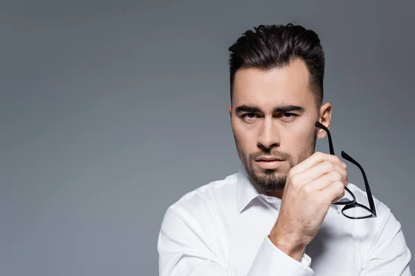 Hombre de negocios barbudo con camisa blanca sosteniendo gafas y mirando a la cámara aislada en gris - foto de stock