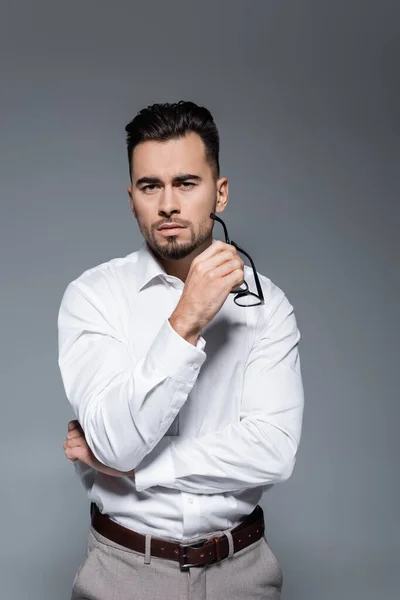 Hombre barbudo con camisa blanca sosteniendo gafas y mirando a la cámara aislada en gris - foto de stock