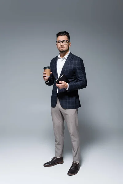 Pleine longueur de l'homme d'affaires dans des lunettes et blazer tenant tasse en papier et bloc-notes sur gris — Photo de stock