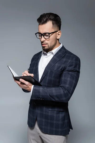 Empresário em blazer e óculos escrevendo em notebook isolado em cinza — Fotografia de Stock