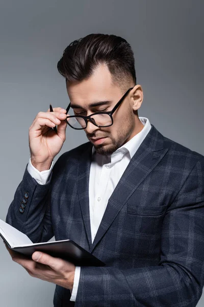 Geschäftsmann in Blazer und Brille blickt auf Notizbuch isoliert auf Grau — Stockfoto