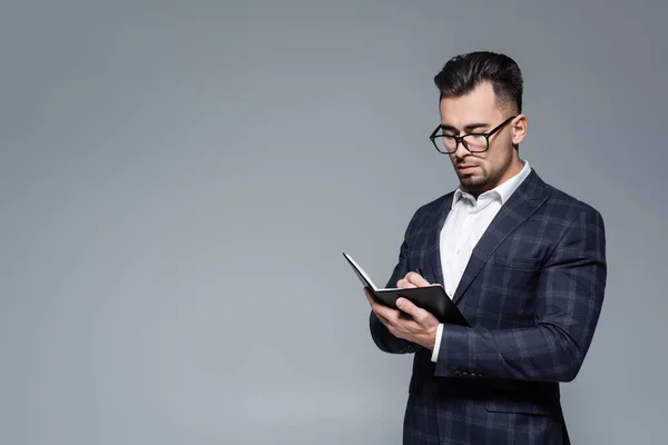 Joven hombre de negocios con traje y gafas escribiendo en cuaderno aislado en gris — Stock Photo