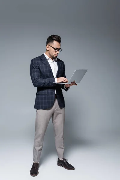 Full length of businessman in suit and glasses using laptop on grey — Stock Photo