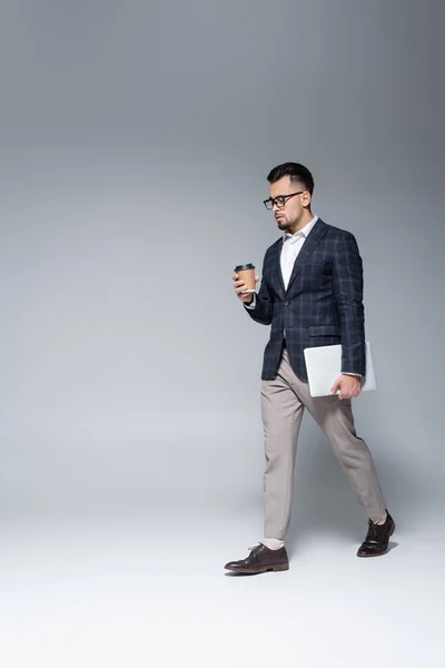 Longitud completa de hombre de negocios en traje y gafas que sostienen el ordenador portátil y caminar con taza de papel en gris - foto de stock