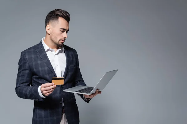 Uomo d'affari in giacca e cravatta in possesso di carta di credito e utilizzando computer portatile isolato su grigio — Foto stock