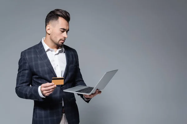 Hombre de negocios en traje con tarjeta de crédito y el uso de ordenador portátil aislado en gris - foto de stock