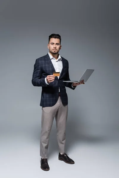 Full length of businessman in suit holding credit card and laptop on grey — Stock Photo