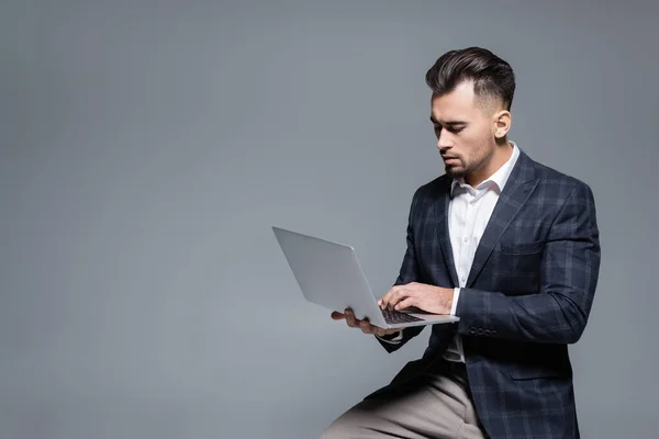 Hombre de negocios barbudo en traje usando portátil aislado en gris - foto de stock