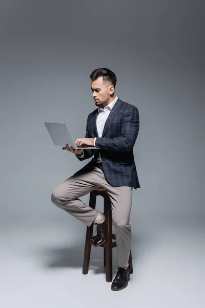 Longitud completa de hombre de negocios barbudo en traje usando el ordenador portátil y sentado en el taburete en gris - foto de stock