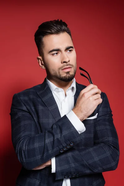 Hombre de negocios barbudo en traje con chaqueta a cuadros sosteniendo gafas en rojo - foto de stock