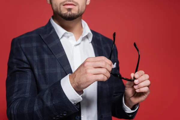 Cropped view of young businessman in checkered blazer holding glasses isolated on red — Stock Photo