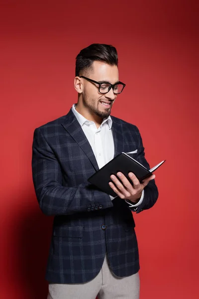 Happy businessman in checkered suit and glasses smiling while writing in notebook on red — Stock Photo