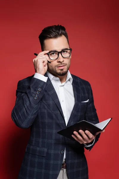 Bearded businessman in checkered suit adjusting glasses while holding pen and notebook on red — Stock Photo