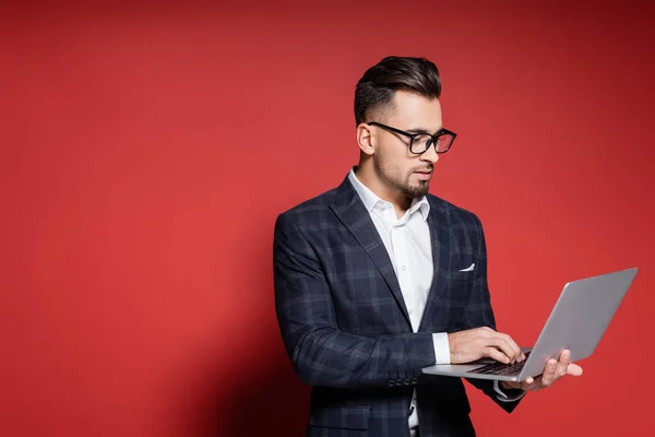 Bearded businessman in checkered blazer and glasses using laptop on red — Stock Photo