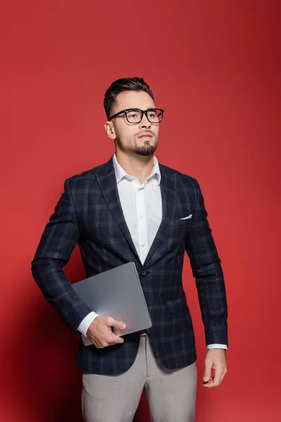 Bearded businessman in blazer and glasses holding laptop on red — Stock Photo