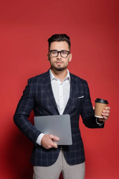 Homme d'affaires barbu en blazer et lunettes tenant ordinateur portable et tasse en papier sur rouge — Photo de stock