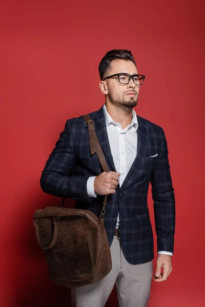 Homme d'affaires barbu en blazer et lunettes debout avec sac en cuir sur rouge — Photo de stock