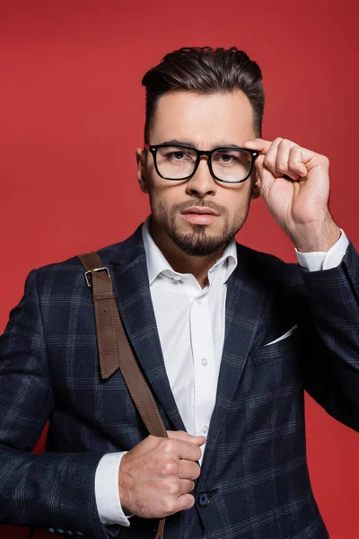 Bearded businessman in blazer adjusting glasses and holding leather bag on red — Stock Photo