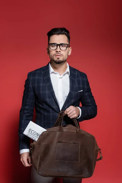 Bearded businessman in blazer and glasses holding leather bag with newspaper on red — Stock Photo