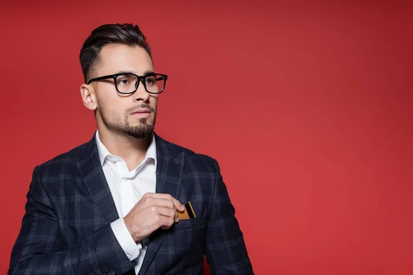 Hombre de negocios barbudo en blazer y gafas poniendo tarjeta de crédito en el bolsillo en rojo - foto de stock